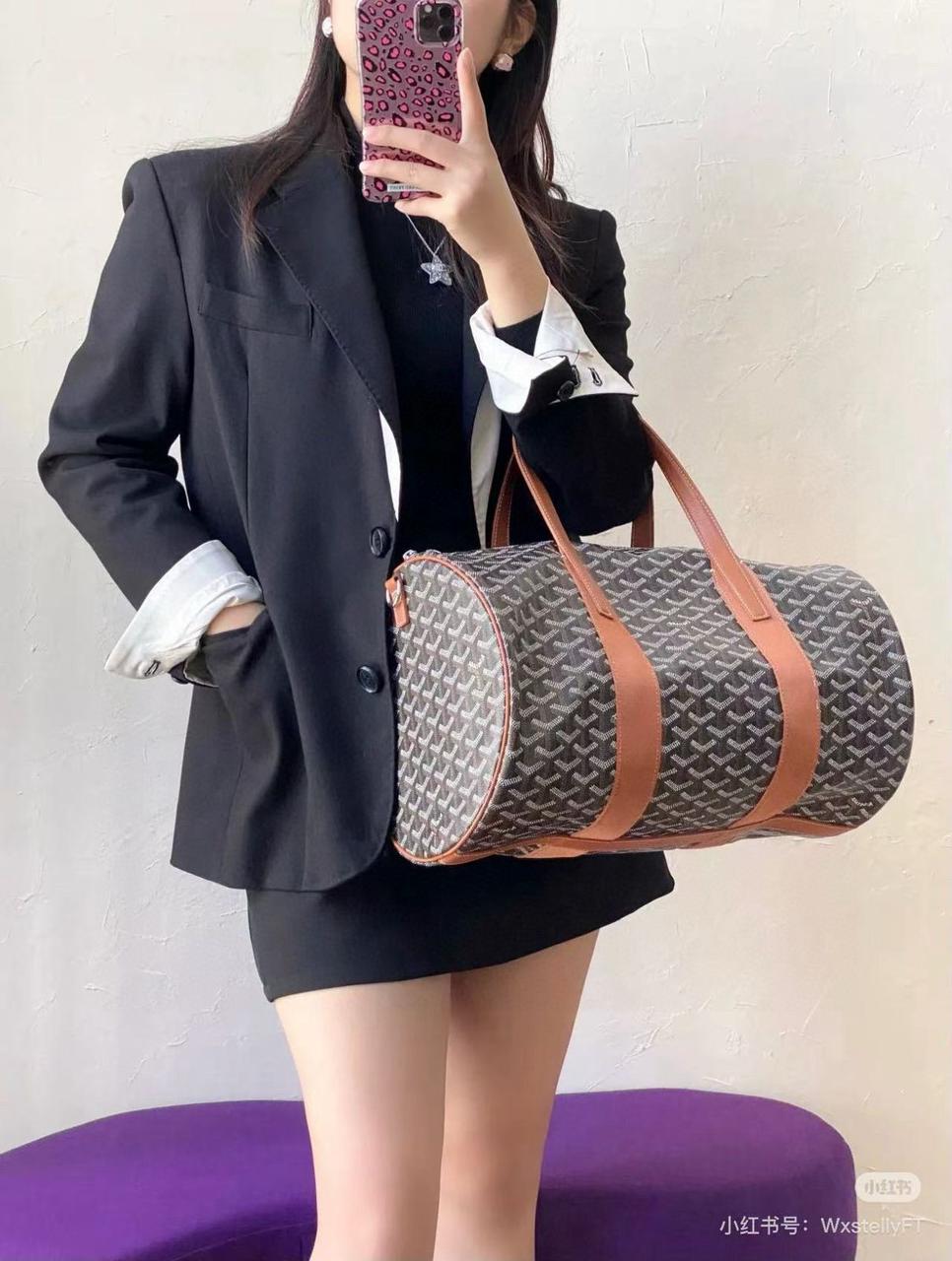 Model in blazer holding black and tan Goyard Goyardine canvas cylinder duffel bag